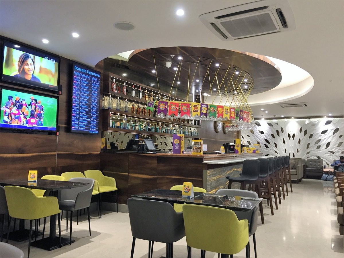 The travel club lounge at Chhatrapati Shivaji Airport, featuring a bar space with an assortment of green, charcoal and black seating options including bar stools. There is a flight monitor on the left hand wall and two televisions.