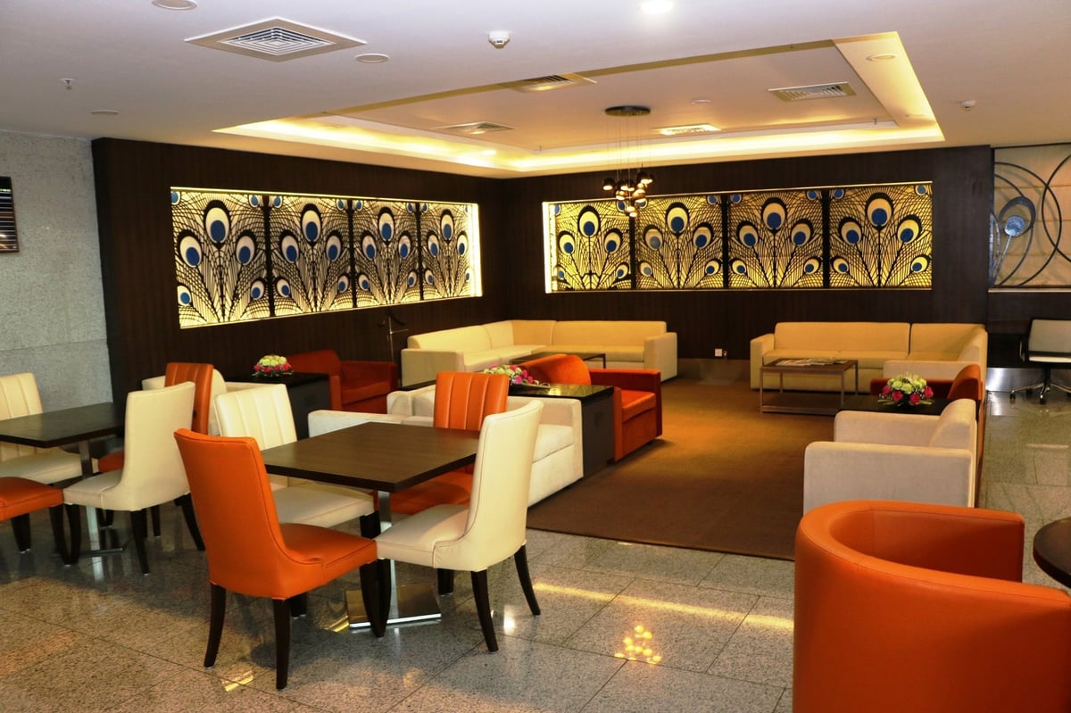 The Aviserv Lounge (west) at t2 of Mumbai Airport featuring orange and beige sofas and dining options with peacock feature artwork on the dark brown back walls. The space is warmly lit and there are flower arrangements on counters.