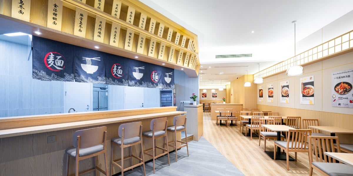 The landslide plaza premium lounge at CKG airport. Featuring a stylish dining area with light wood furnishings. There is a bar style counter with wooden stalls in front of a kitchen, partially covered by traditional Japanese-style noren curtains.