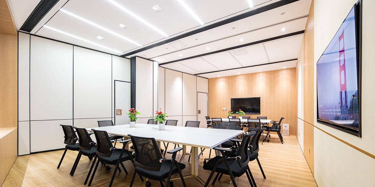 The image shows a modern conference room at the Plaza Premium Lounge Landside at CKG Airport. It has a clean, minimalist design. The room features a long white tables surrounded by black mesh office chairs on wheels. The floor is made of light wood.