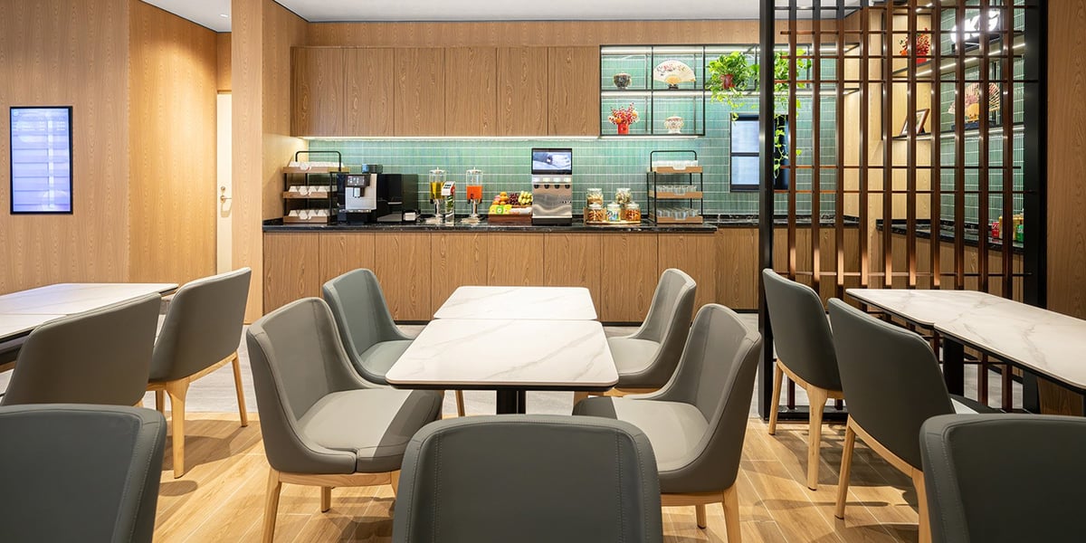 The Plaza Premium Lounge at CKG, pier H. It features a modern dining area with marble top tables, upholstered grey chairs, and wood panelled walls. The self serve refreshment counter offers a selection of beverages, snacks and fruit.