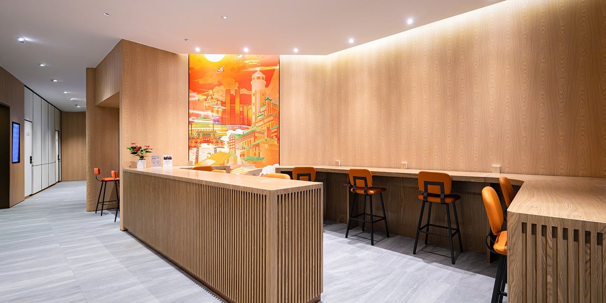 This image shows an area of the Plaza Premium Lounge at CKG airport landside. The area features light wood panelling throughout and at the centre is a counter with vertical wooden slats paired with high bar-style seating. On the back wall is a mural.