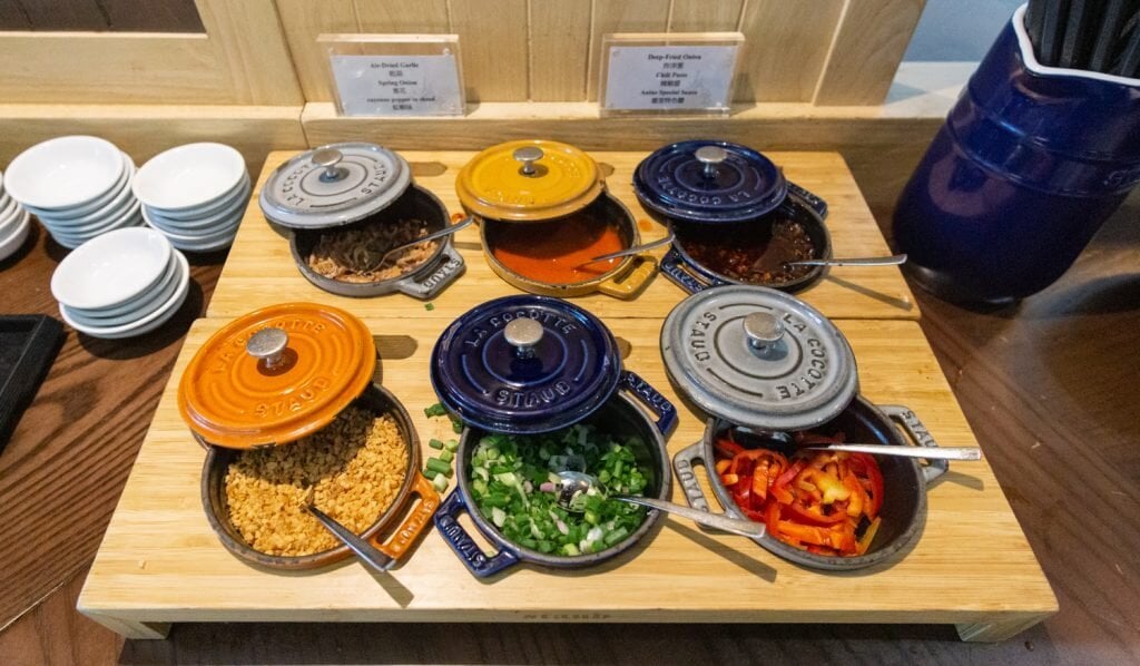 Condiment and toppings station at Club Autus Lounge in Hong Kong International Airport, showcasing colorful Staub mini cocottes filled with spring onions, chili oil, fried garlic, and peppers, arranged beside noodle dishes and soup options.