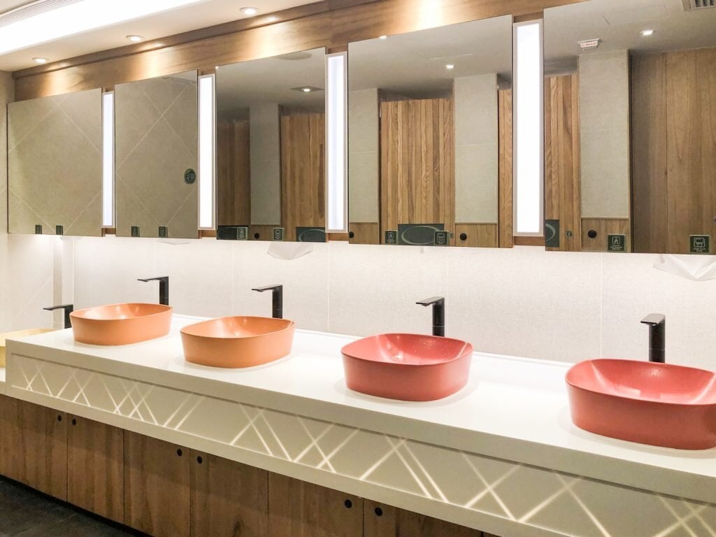 Modern restroom at Club Autus Lounge in Hong Kong International Airport, featuring stylish coral-toned vessel sinks, minimalist black faucets, well-lit vanity mirrors, and wood-accented cabinetry.