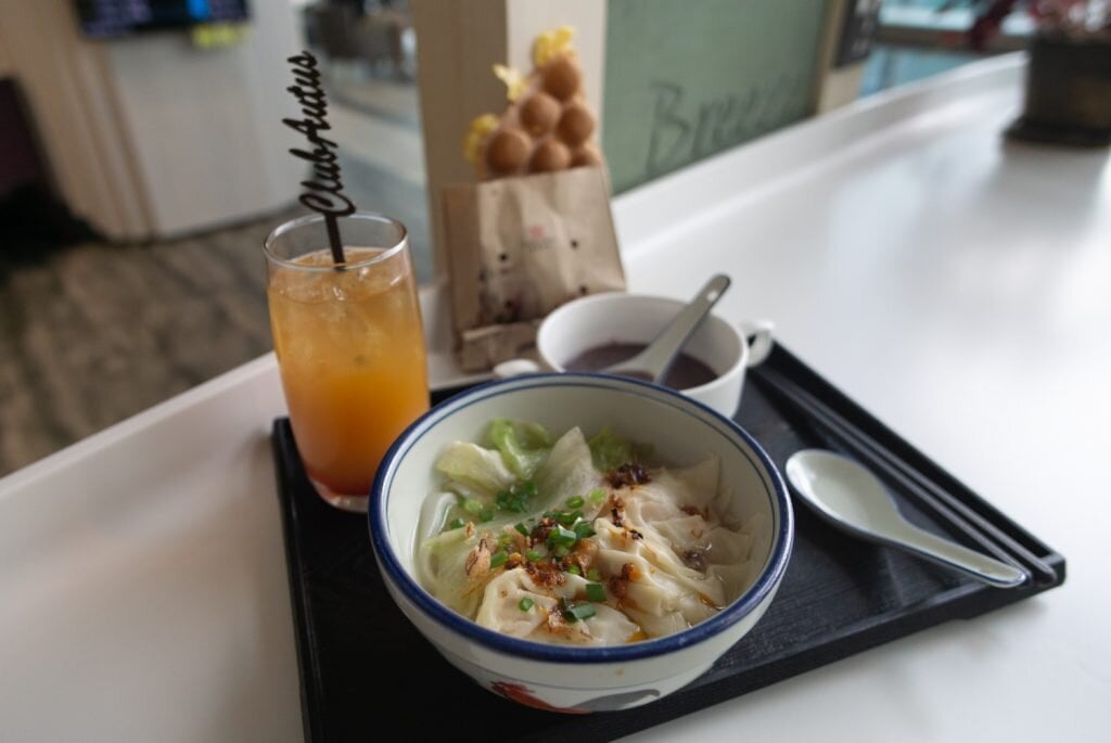Asian meal set at Club Autus Lounge in Hong Kong International Airport, featuring a bowl of wonton noodle soup, iced tea with a branded stirrer, egg waffles, and a dessert soup, served on a tray at the dining area.