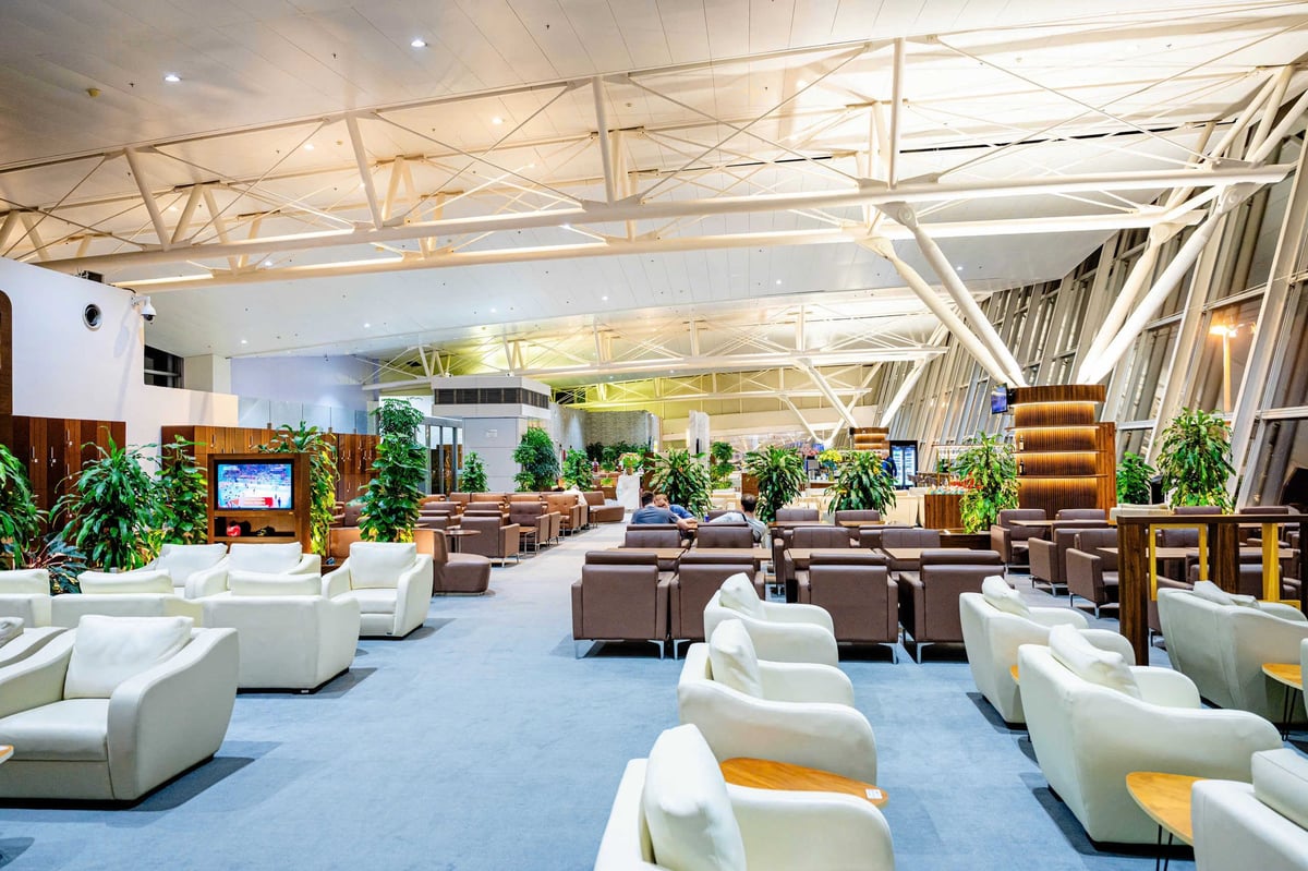 Spacious seating area at the ACV Business Lounge, Hanoi Airport. The lounge features a combination of white and brown leather armchairs arranged in rows with small wooden side tables. Tall indoor plants and a modern design with high ceilings