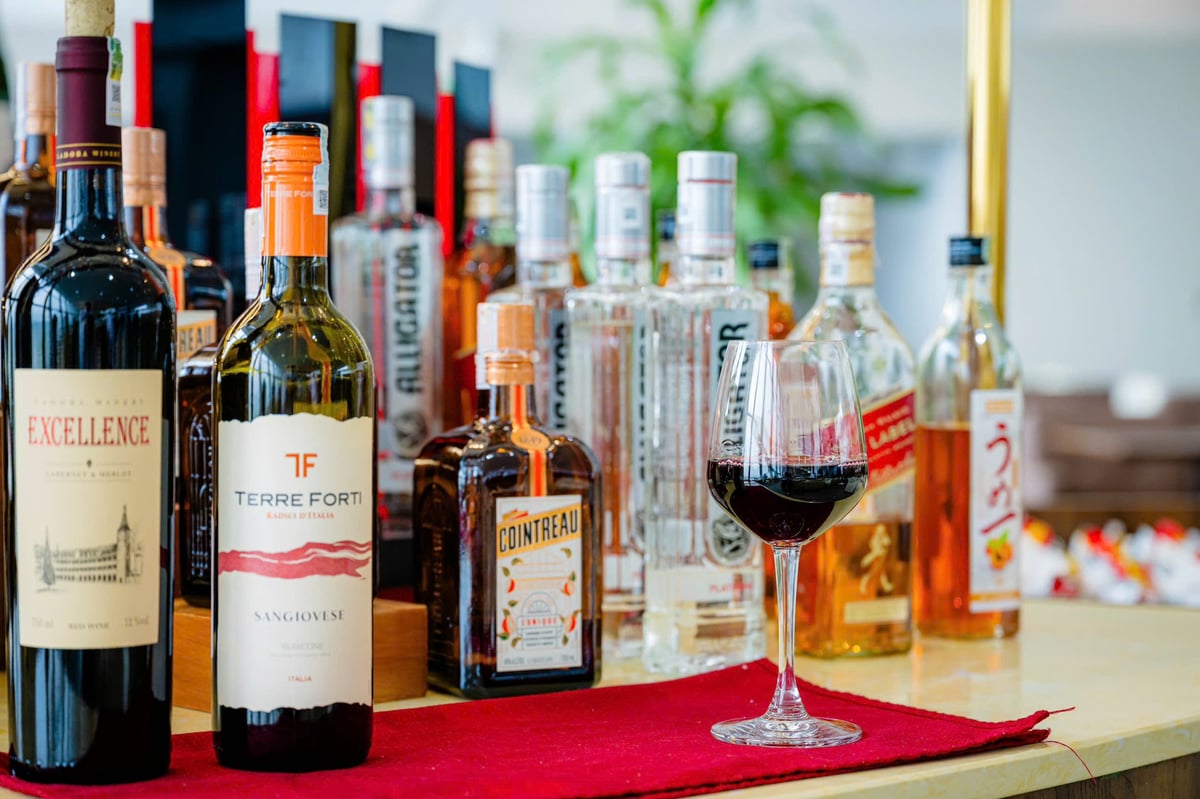 Selection of alcoholic beverages at ACV Business Lounge, Hanoi Airport. A glass of red wine sits on a red mat in front of various bottles including Terre Forti Sangiovese, Cointreau, and other premium spirits, creating an inviting bar setup.