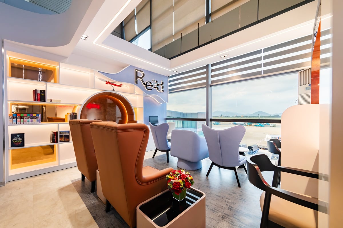 Reading lounge area at ASG Sky Lounge, Noi Bai Domestic Airport, featuring modern leather and fabric armchairs, a well-lit bookshelf with books and magazines, and panoramic windows with runway and mountain views