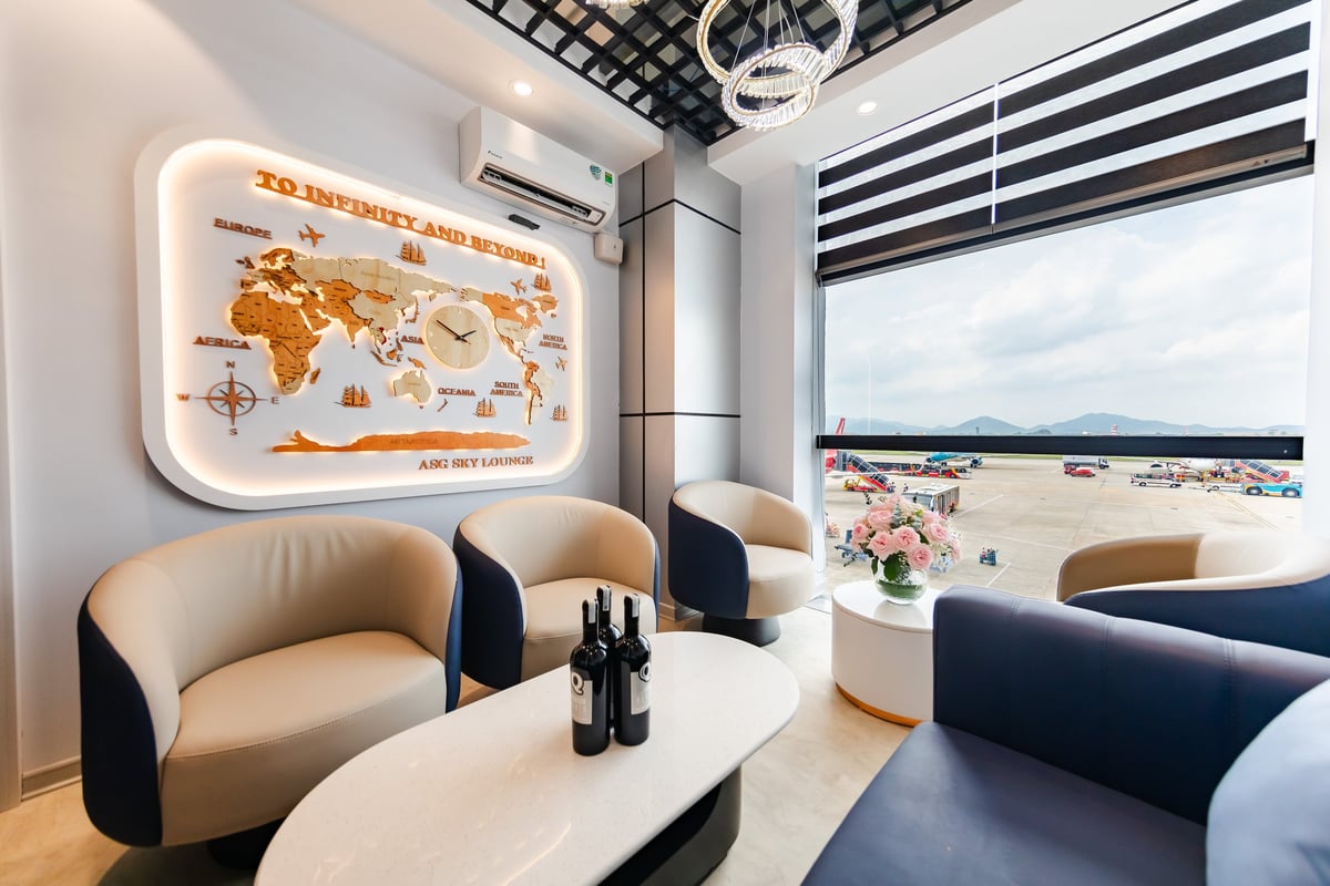 ASG Sky Lounge at Hanoi Airport, featuring a cozy seating corner with modern cream and navy armchairs, a white marble coffee table with wine bottles, and a decorative wall map with a central clock that reads 