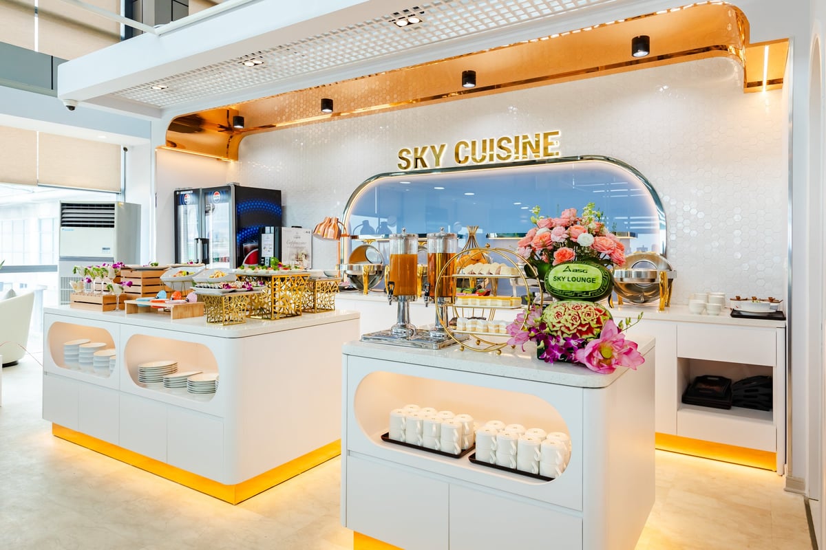 Buffet area at ASG Sky Lounge, Noi Bai Domestic Airport, featuring elegant white counters with golden accents, fresh flower arrangements, juice dispensers, pastries, salads, and a variety of hot and cold dishes under the sign 