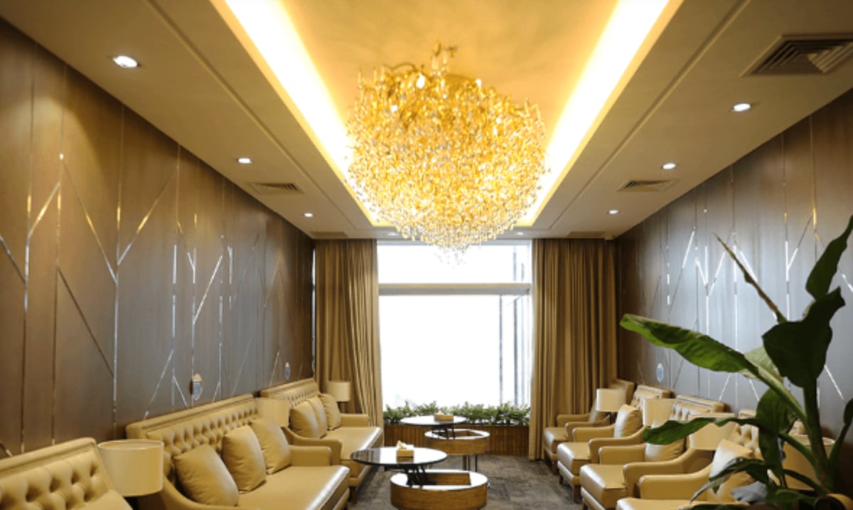 Luxurious seating area at the Bamboo Airways First Lounge in Hanoi Airport, featuring cream-colored leather sofas and armchairs, elegant round wooden coffee tables, gold-toned walls, and a statement chandelier under warm ambient lighting.