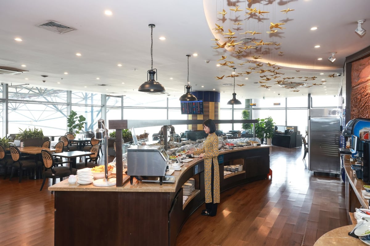 Buffet and dining area at SH Premium Lounge in Hanoi Airport Terminal 1, with a guest serving food, modern pendant lighting, wooden flooring, decorative ceiling art featuring golden birds, and large windows providing natural light.