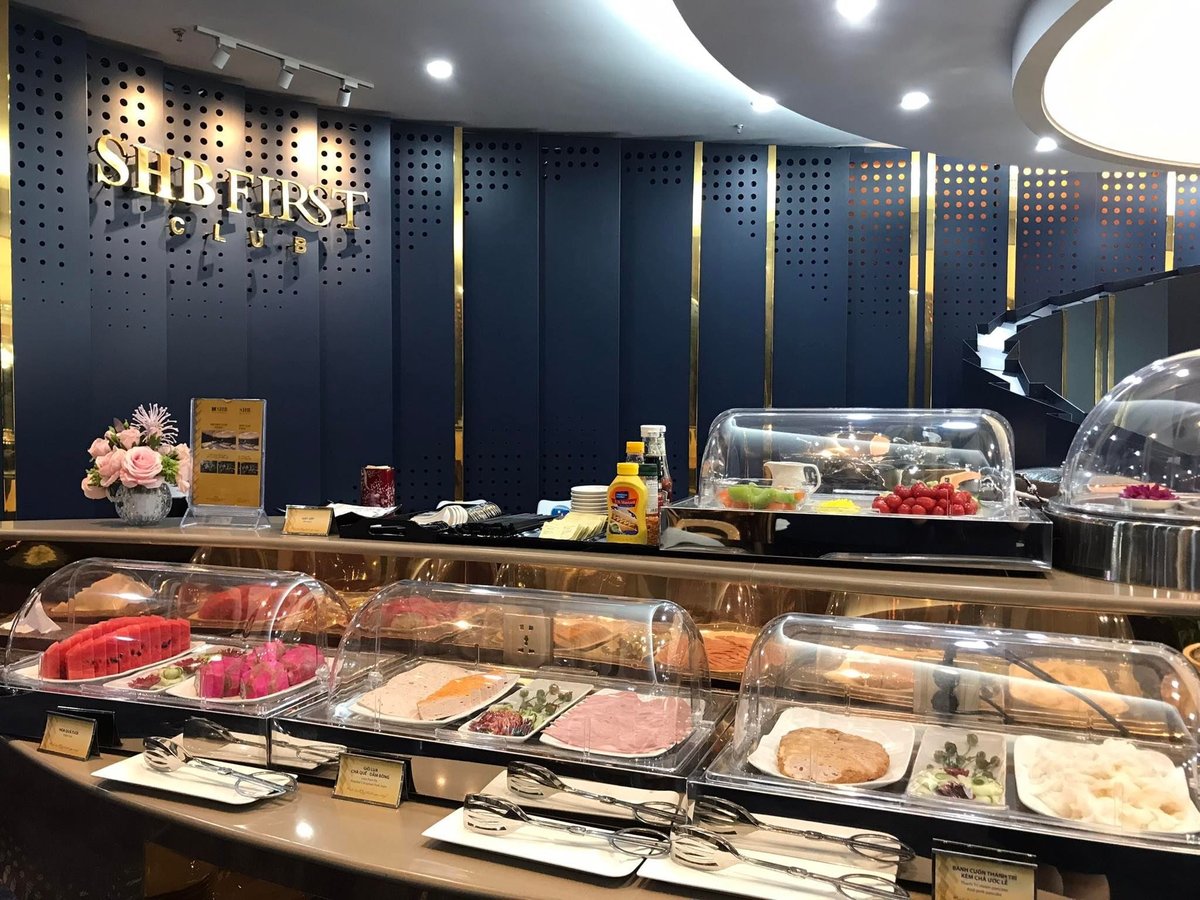 Buffet spread at SHB First Club Lounge in Hanoi Airport, featuring a selection of cold cuts, fresh fruits like watermelon and dragon fruit, assorted pastries, and condiments, all neatly arranged in covered trays.