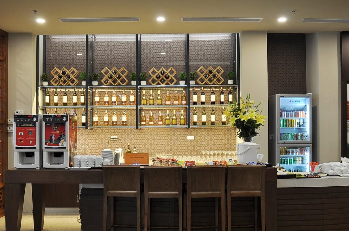 Buffet and drink station at the Vietnam Airlines Lotus Lounge in Hanoi Airport. The area features a counter with wooden bar stools, two self-service coffee machines, and a variety of snacks neatly arranged.