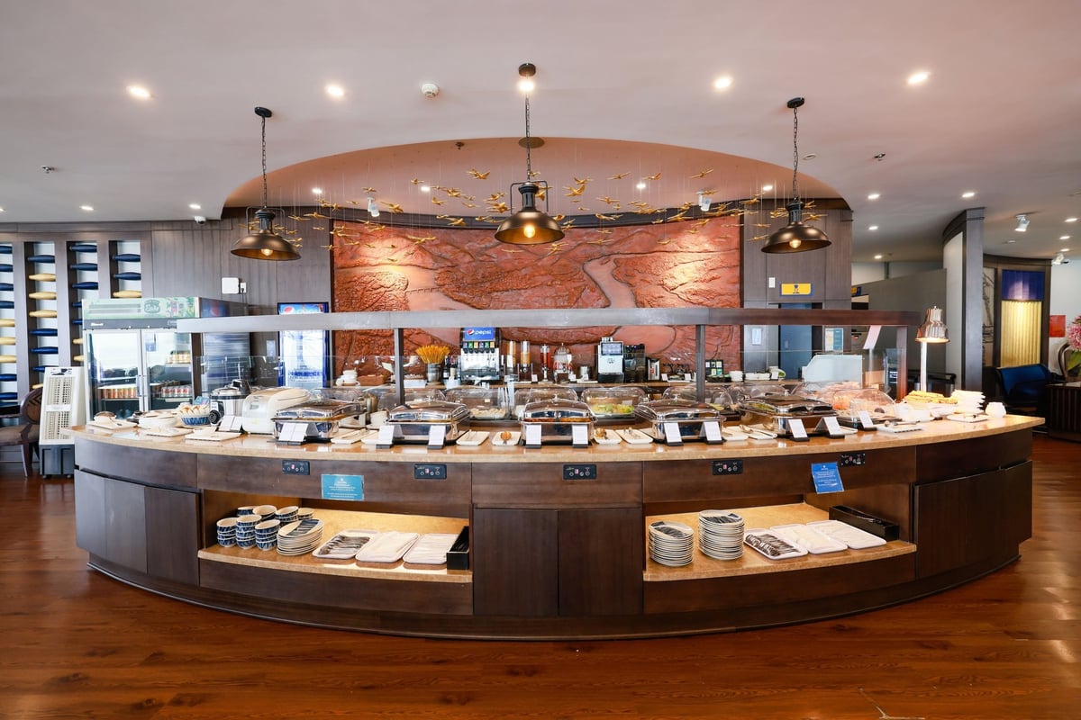Buffet counter at the SH Premium Lounge in Hanoi Airport Terminal 1, showcasing a wide variety of hot dishes, salads, and beverages, with elegant wooden cabinetry, hanging pendant lights, and a decorative wall map backdrop.