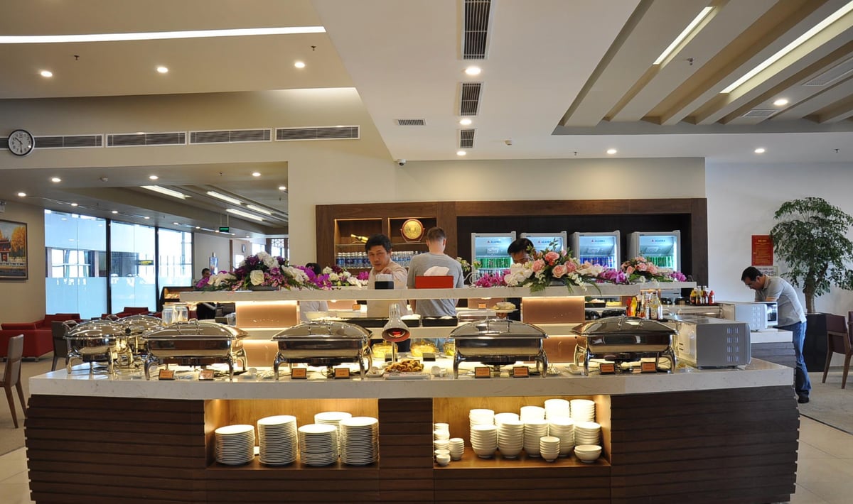 Buffet area at the Vietnam Airlines Lotus Lounge in Hanoi Airport, showcasing a selection of chafing dishes filled with hot food. The counter features neatly stacked plates and bowls on the lower shelves.