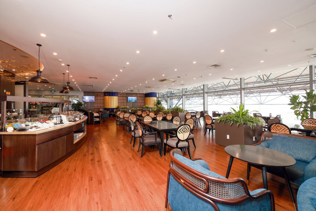 Spacious dining area at SH Premium Lounge in Hanoi Airport Terminal 1, featuring elegant wooden tables, patterned chairs, indoor plants, large windows with runway views, and a buffet counter along the side.