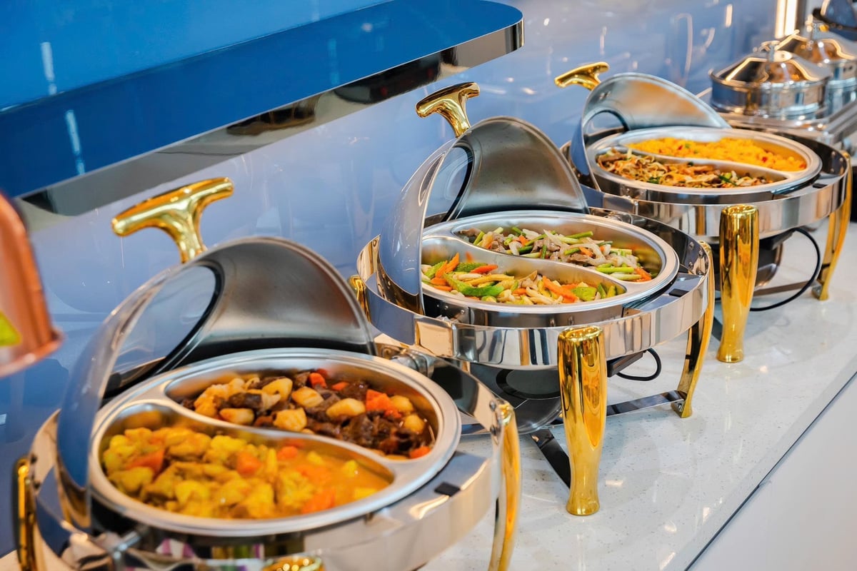 Close-up of a hot buffet station at ASG Sky Lounge, Hanoi Airport, showcasing stainless steel and gold chafing dishes filled with colorful stir-fried vegetables, curries, rice, and noodle dishes.
