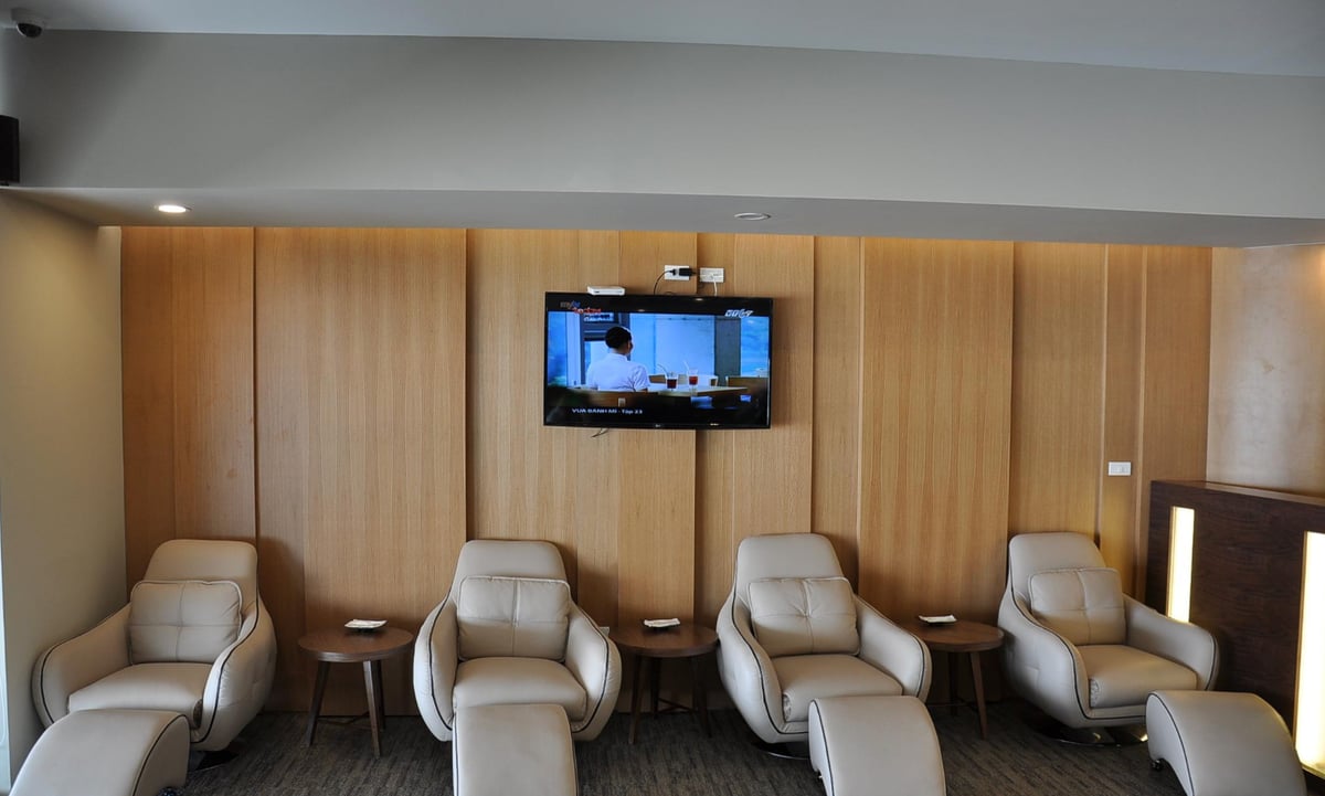 Massage chair area at the Vietnam Airlines Lotus Lounge in Hanoi Airport. The space features a row of plush beige leather recliner chairs with footrests, positioned in front of a wall-mounted TV displaying a program.