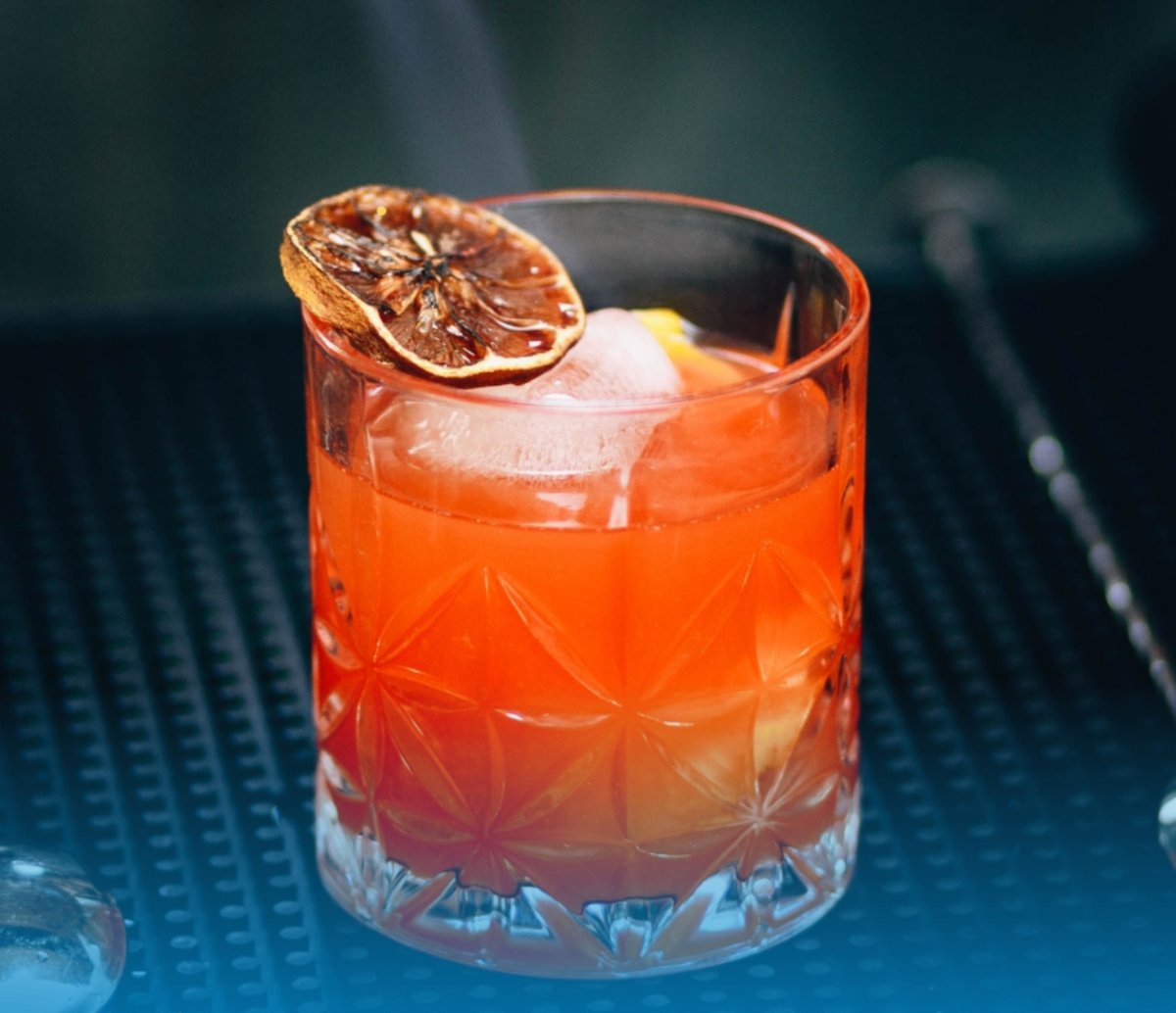 Close-up of a vibrant orange-red cocktail served in a cut-glass tumbler with large ice cubes and garnished with a dried citrus slice on top.