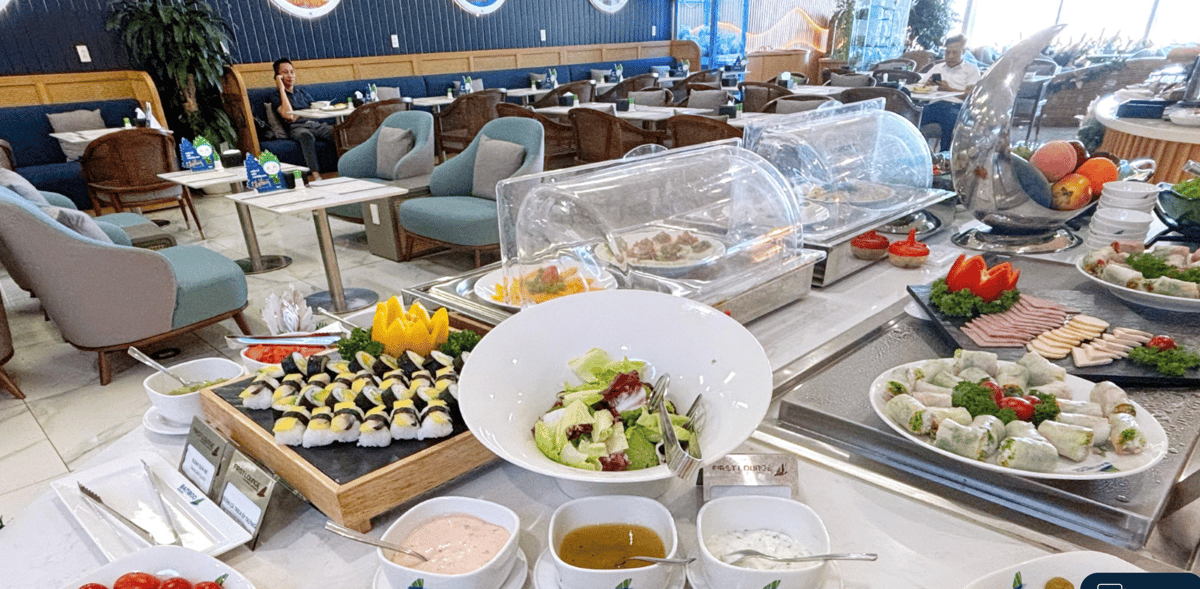 Buffet spread at the Bamboo Airways First Lounge in Hanoi Airport, featuring a variety of fresh dishes including spring rolls, cold cuts, fruits, and salads, displayed on a round white counter.