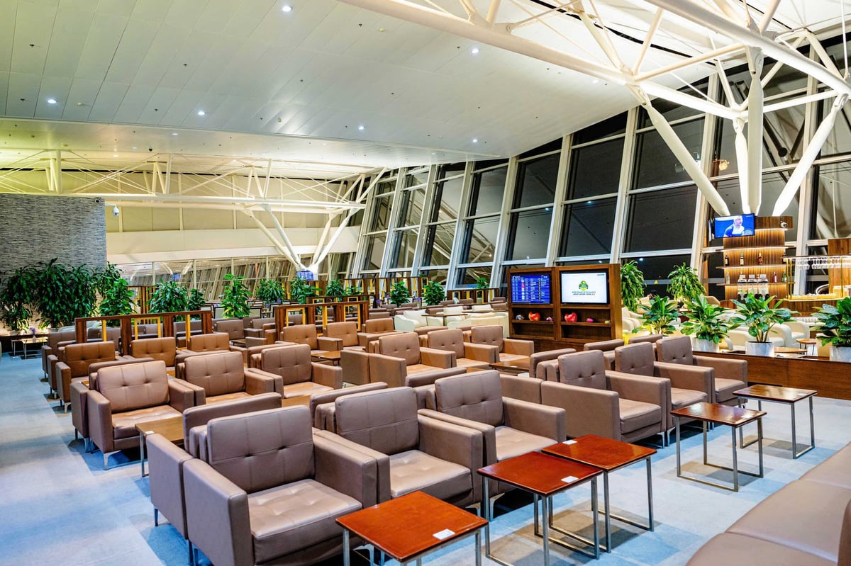 Seating area of the ACV Business Lounge at Hanoi Noi Bai International Airport. The lounge features rows of comfortable brown armchairs with wooden side tables, complemented by green potted plants and large windows providing natural light.