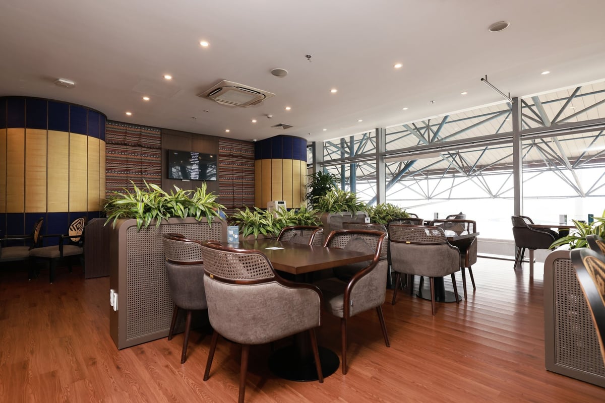 Elegant seating area at SH Premium Lounge, Hanoi Airport Terminal 1, featuring wooden floors, cushioned chairs with rattan backs, indoor greenery, large windows with tarmac views, and modern ceiling lighting.