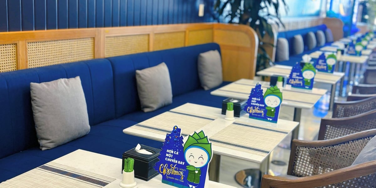 Close-up of the dining area at the Bamboo Airways First Lounge in Hanoi Airport, showcasing navy-blue cushioned seating with gray accent pillows, modern rattan-back chairs, and neatly set tables with placemats and tissue boxes.