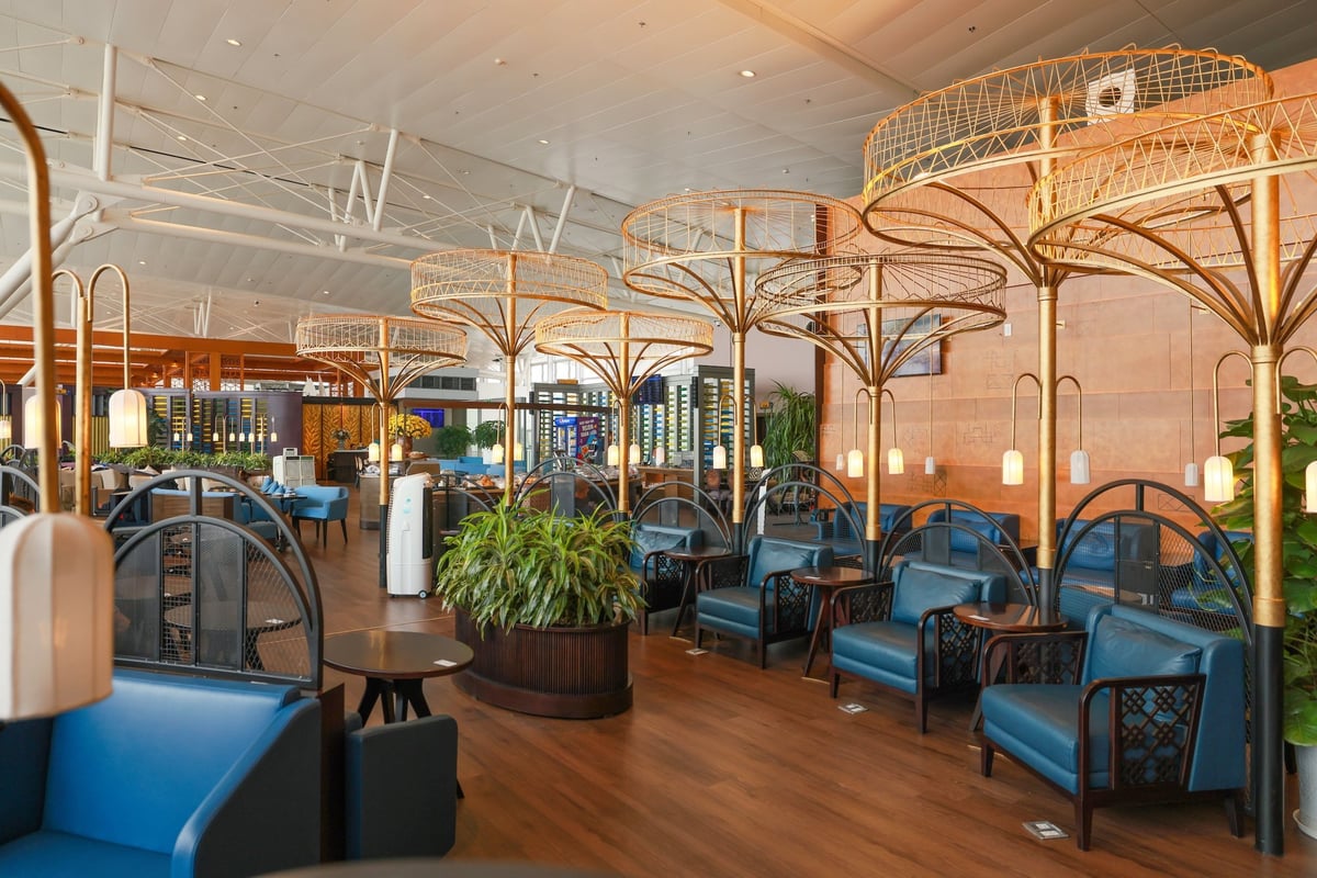 Seating area of the SH Premium Lounge at Hanoi International Airport, featuring stylish blue armchairs with wooden armrests arranged under golden tree-like light fixtures.