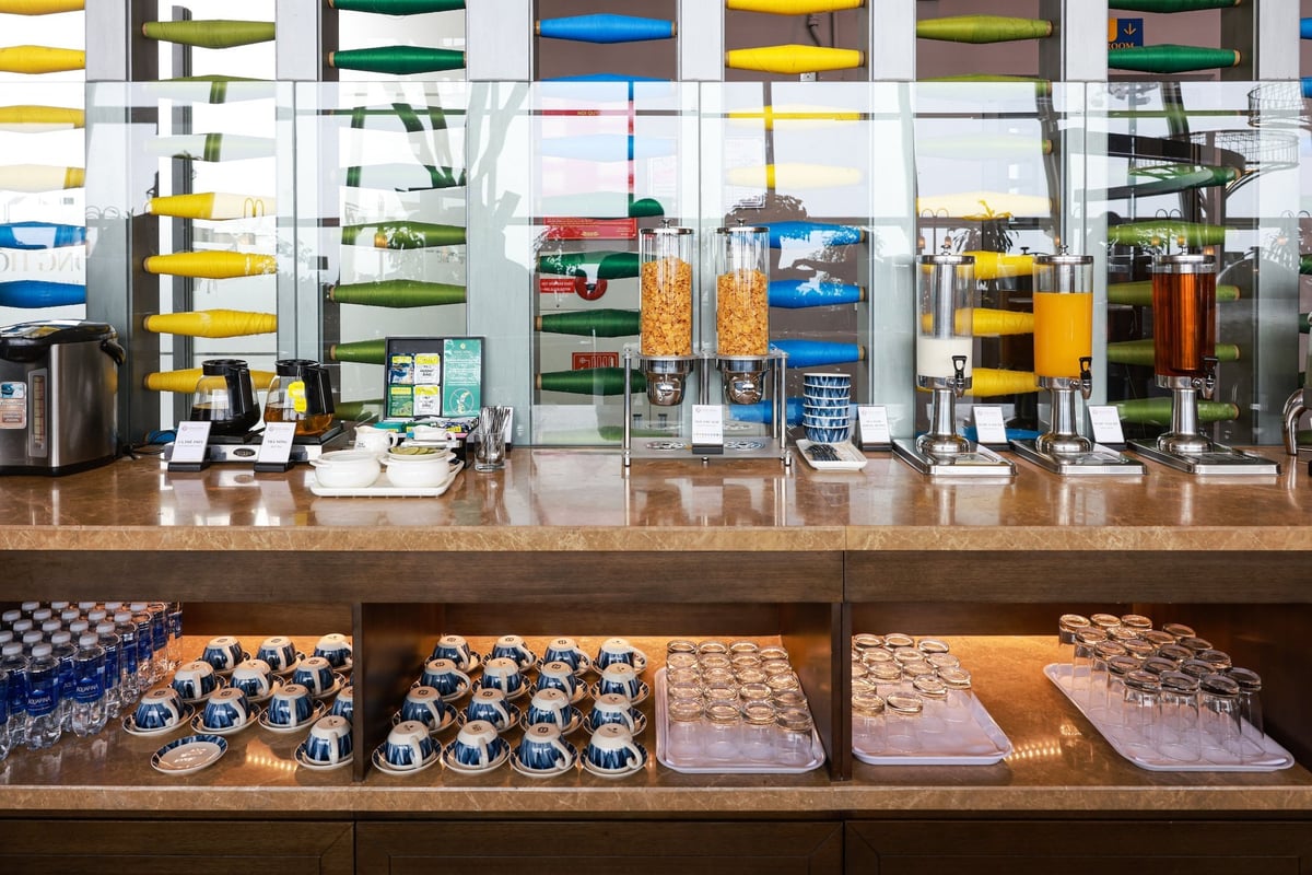 Beverage and cereal station at the SH Premium Lounge, Hanoi Airport, featuring dispensers with cornflakes, milk, orange juice, and iced tea. Below the counter are neatly arranged glasses, cups, and bottled water.