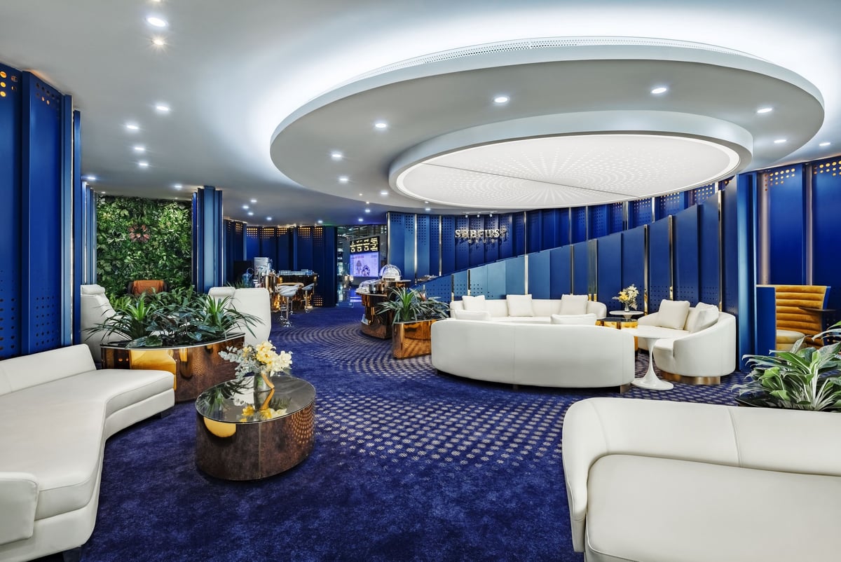 Luxurious SHB First Club Lounge at Hanoi Airport featuring modern design with white leather sofas, rich blue carpeting, and gold accents. The space is highlighted by a striking circular ceiling light, and a green vertical garden wall.