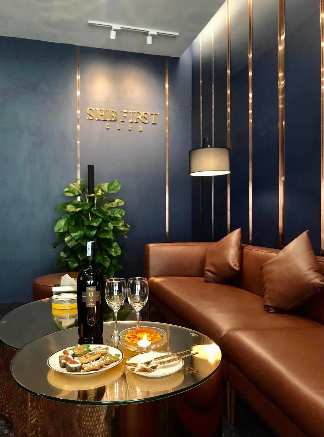 Cozy seating area at the SHB First Club Lounge in Hanoi Airport, featuring a brown leather sofa with matching cushions, a sleek glass coffee table set with a bottle of red wine, two wine glasses, plates of snacks, and a lit tealight candle.