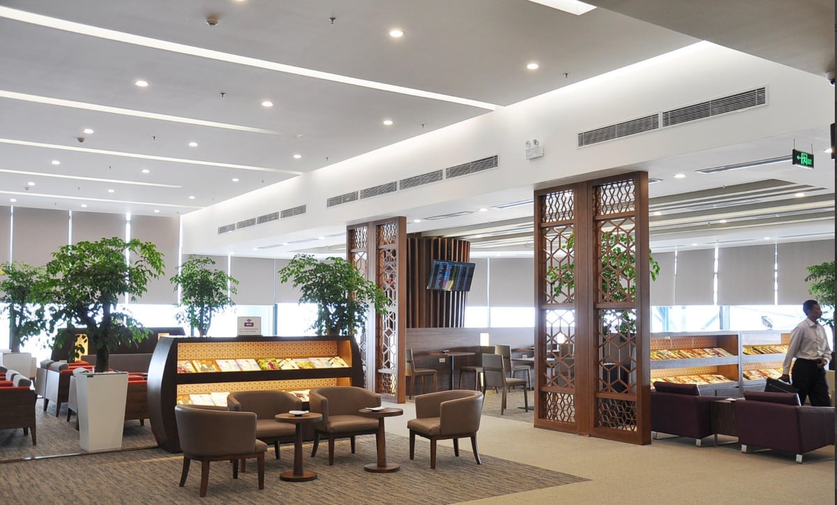 Spacious seating area at the T1 Vietnam Airlines Lotus Lounge at HAN airport, featuring a mix of brown armchairs and wooden tables. A backlit magazine rack is centrally placed, and potted plants add a natural touch.