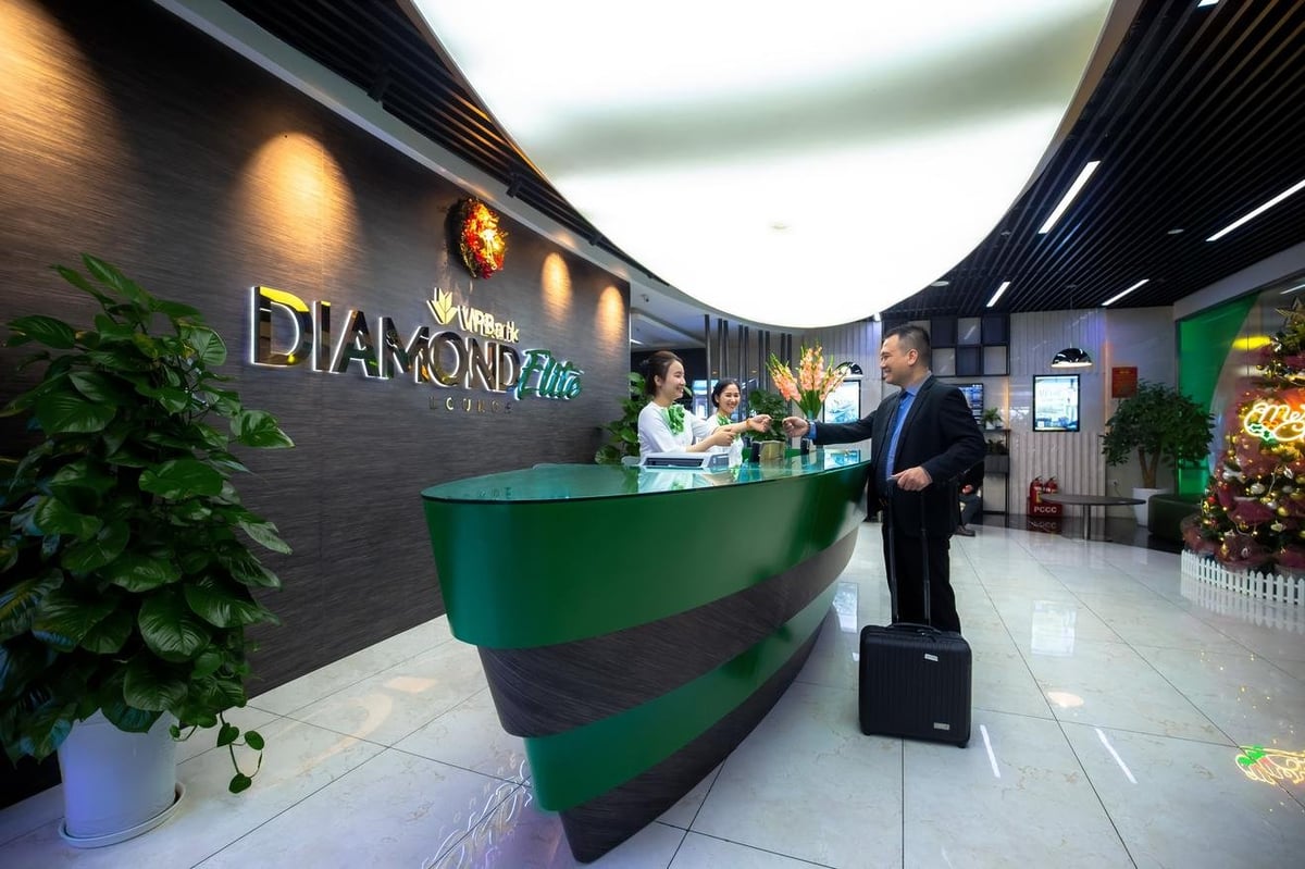 Reception area of the VPBank Diamond Elite Lounge at Hanoi Airport, featuring a sleek modern design with a green and dark wood reception desk. Two friendly staff members are assisting a male guest with a suitcase.