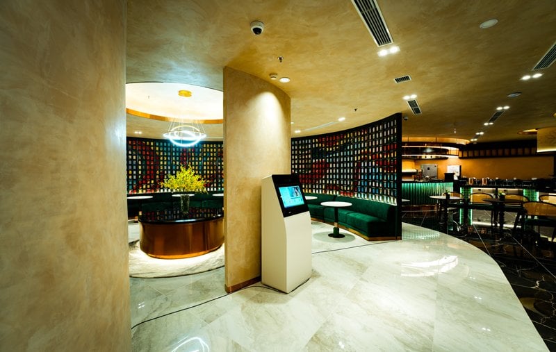 Interior of the VPBank Diamond Elite Lounge at Hanoi Airport, showcasing a sophisticated design with warm beige walls, marble flooring, and modern lighting. A digital check-in kiosk is placed near a round table with floral arrangements