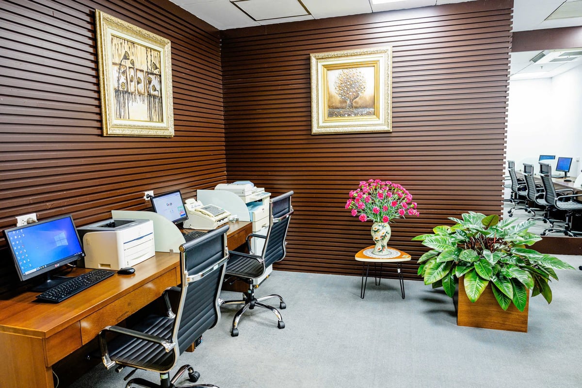Workstation area at ACV Business Lounge, Hanoi Airport, featuring desktop computers, printers, and fax machines on a wooden desk with black swivel chairs. Decorative elements include framed artwork, a vase of pink flowers, and potted greenery.