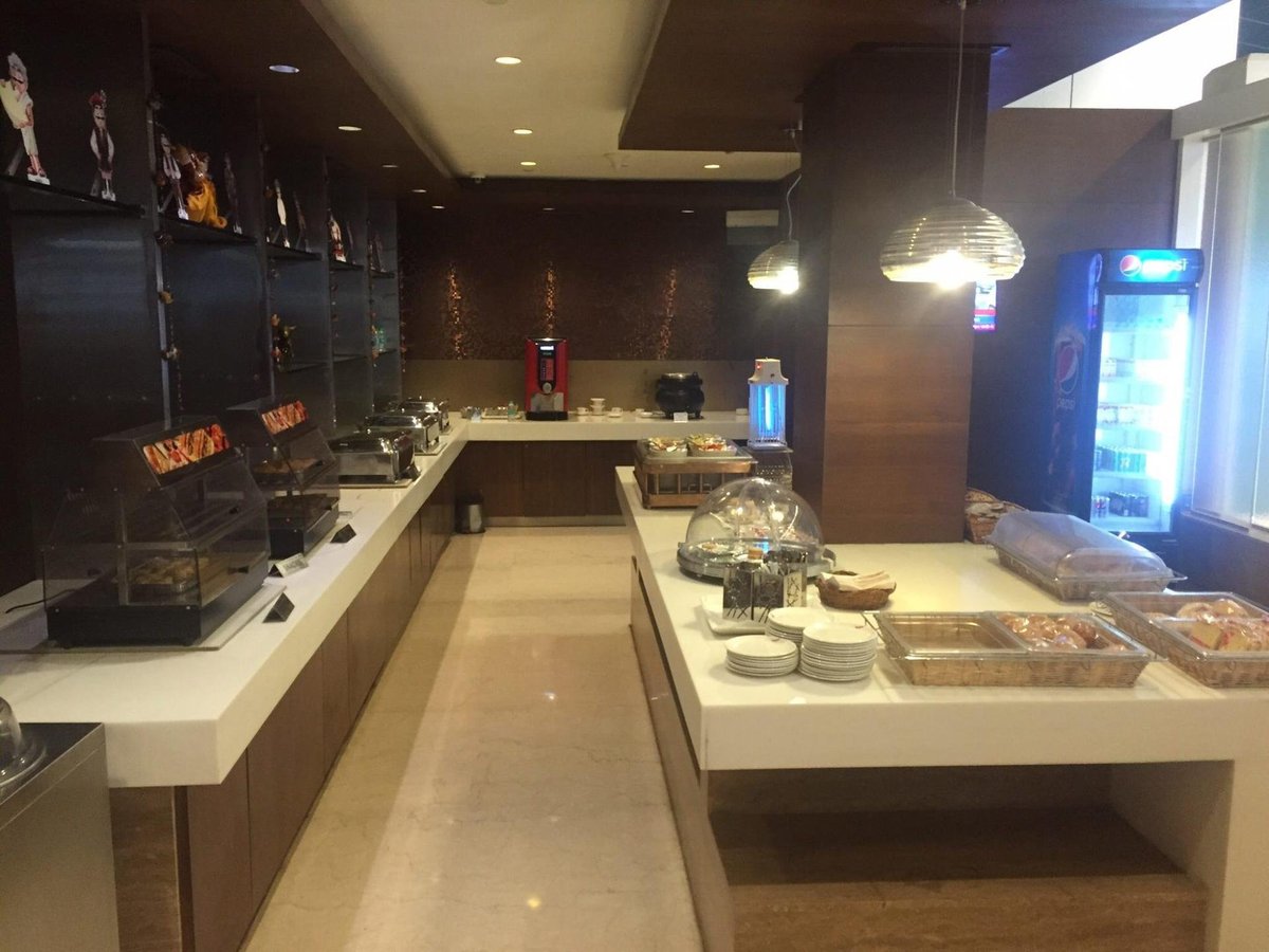 Buffet Area inside airport lounge, features a selection of hot and cold foods displayed under protective covers. The counter is stocked with plates and cutlery. Backdrop includes a beverage dispenser, and the decor is accentuated with shelved art.