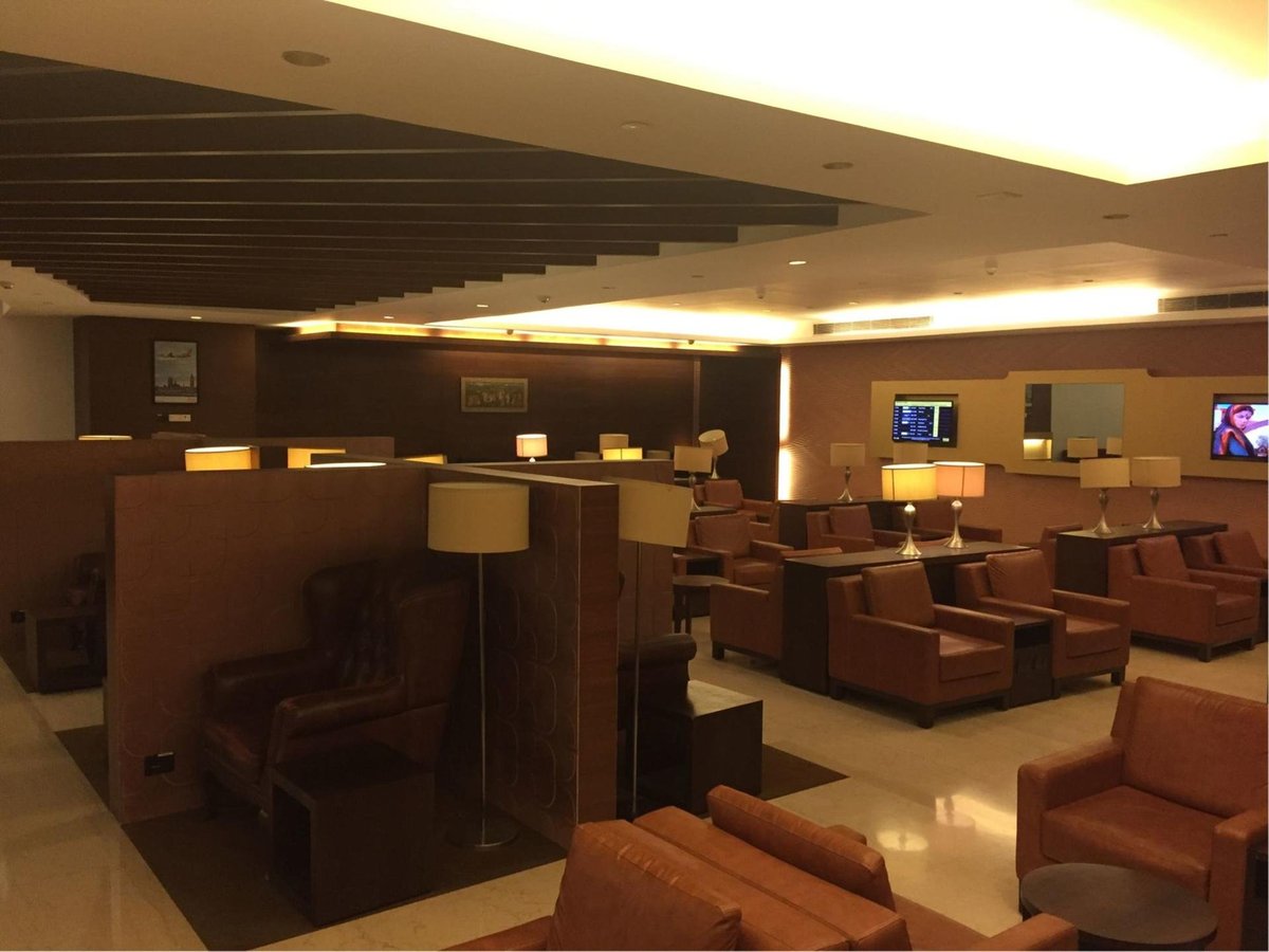 Interior of airport lounge with a warm inviting ambiance, showcasing tan leather armchairs and booths. Ambient lighting from lamps and ceiling fixtures cast a soft glow, while flight information screens provide updates for travellers.
