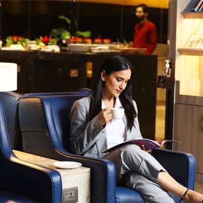A woman in a business suit seated in a blue leather chair in airport lougnge, enjoying a cup of coffee while reading a magazine.  The lounge has a sophisticated ambiance and warm lighting, with a selection of refreshments in the background.