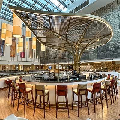 A modern airport lounge with a striking central feature resembling a tree canopy, topped with a mesh screen. The bar is surrounded by high stools, and the area illuminated by natural light filtering through a glass atrium. Chic and stylish atmosphere