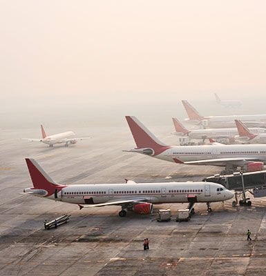 Aerial view of several airplanes with red tails parked on tarmac at dawn, with a hazy sky suggesting mist. Ground crew can be seen working around aircraft, indicating airport ground operations in progress.