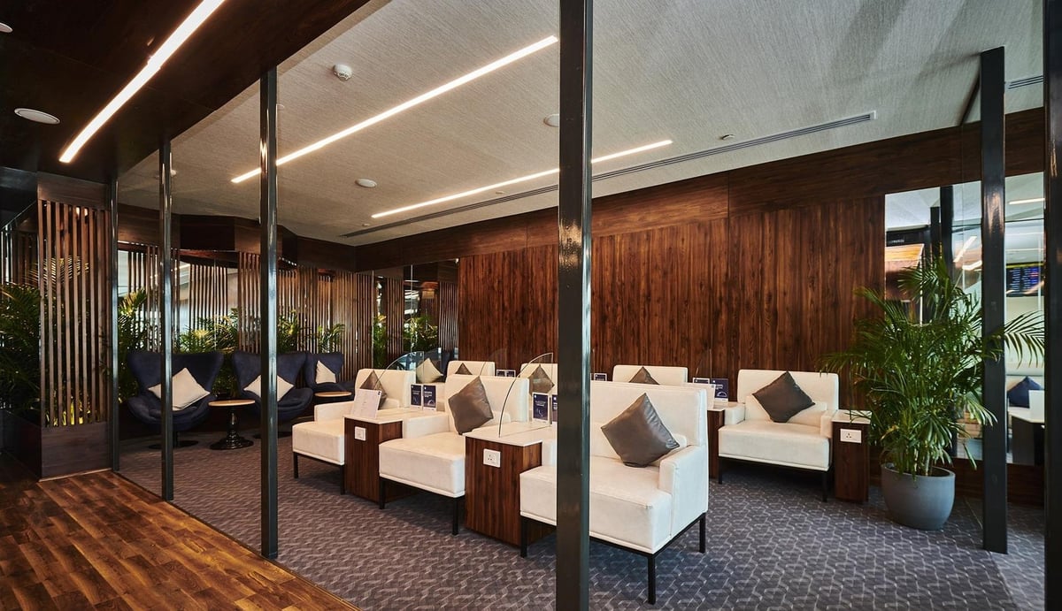 Modern airport lounge featuring wood wall panelling and sleek furniture. Space is divided with vertical wooden slats, creating semi-private seating areas, with plush blue and cream armchairs. Overhead warm lighting complements the natural light.
