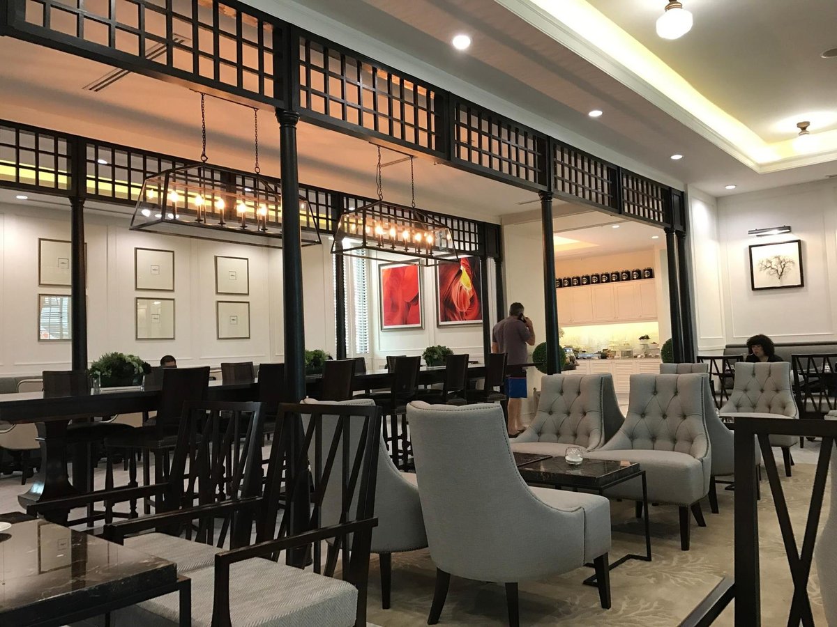 Coral executive lounge at terminal 2 Don Mueang international airport. A blend of classic and contemporary design with tufted grey armchairs, wooden dining tables, and ornate overhead lighting. The room features framed artwork, and a buffet station.