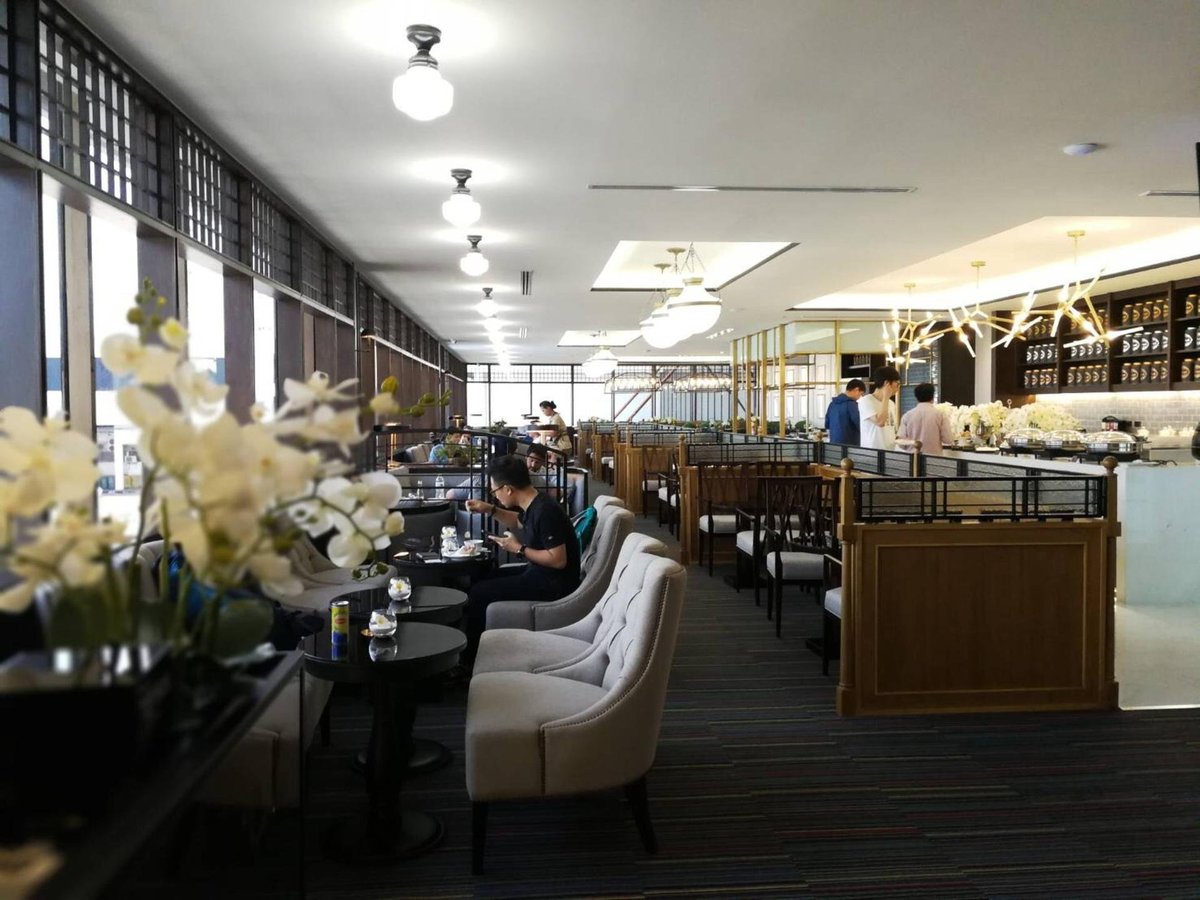 Coral Executive Lounge in terminal 1 of Don Mueang International Airport. A contemporary airport lounge with plush seating and a bright airy ambiance. Comfortable beige sofas lined up beside sleek tables. Buffet and bar area in background with staff.