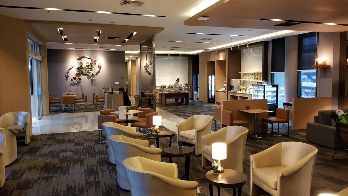Spacious miracle lounge at terminal 2 Don Mueang International Airport with a variety of seating options, including armchairs and dining tables, illuminated by ambient lighting. The back wall is adorned with an artistic metal sculpture.