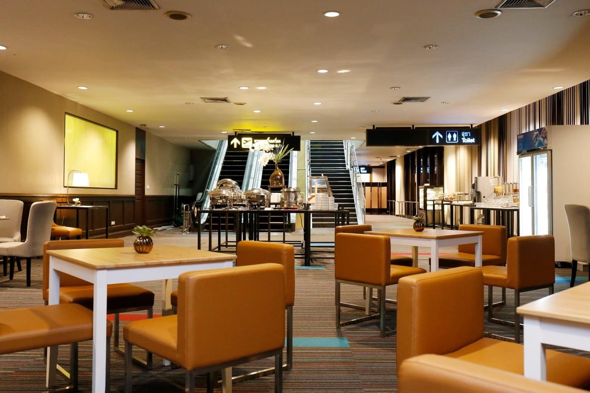 Miracle Lounge No1 in terminal 1 of Don Mueang international airport. Modern lounge with a relaxed and cozy atmosphere. The foreground shows square tables with caramel-colored leather chairs, while the background features a buffet area.