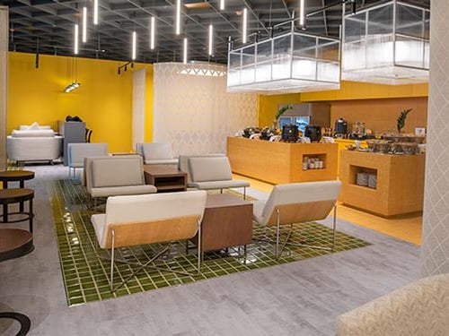 Nok Air Lounge at Don Mueang International Airport. Modern lounge with vibrant yellow walls and a mosaic tiled floor. A self service buffet area positioned on the right. Seating area features contemporary furniture with cream and taupe armchairs.