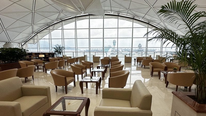 Spacious seating area inside the Emirates Lounge at Hong Kong International Airport, featuring beige armchairs, glass tables, potted plants, and panoramic runway views through floor-to-ceiling windows.