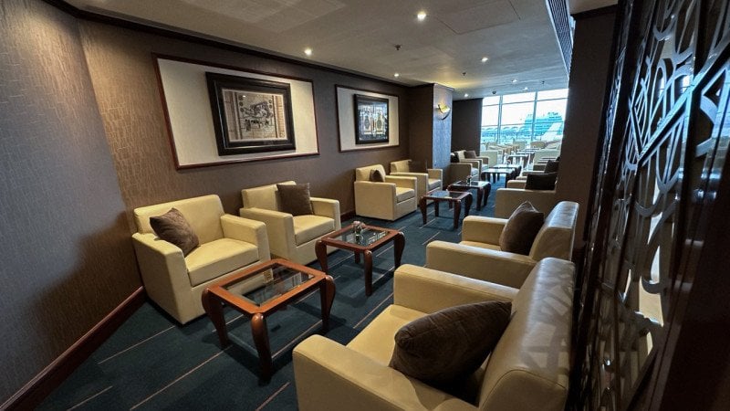 Interior seating area of the Emirates Lounge at Hong Kong International Airport, showcasing cream leather armchairs, dark wood coffee tables, framed artwork, and a carpeted floor with natural light from the windows in the background.