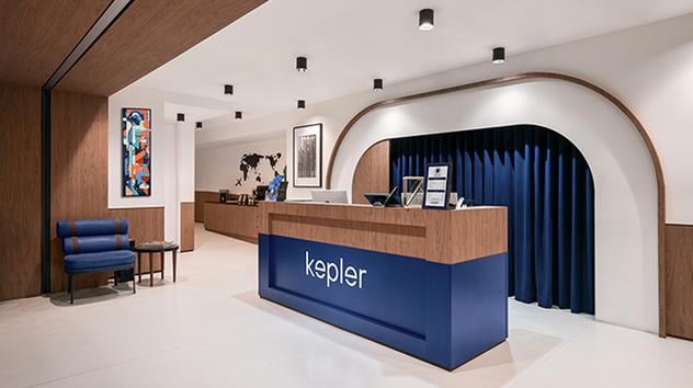 Entrance to the Kepler Club at the Main Terminal Area at Kuala Lumpur Airport featuring a reception desk with blue and wooden accents, a blue curtain behind it, and a blue waiting chair on the left wall. There is also contemporary wall art.
